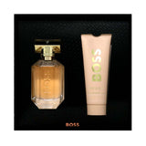 BOSS The Scent cologne set featuring a faceted glass bottle filled with amber liquid and a matching light beige tube, both with gold BOSS branding, set against a black textured background.